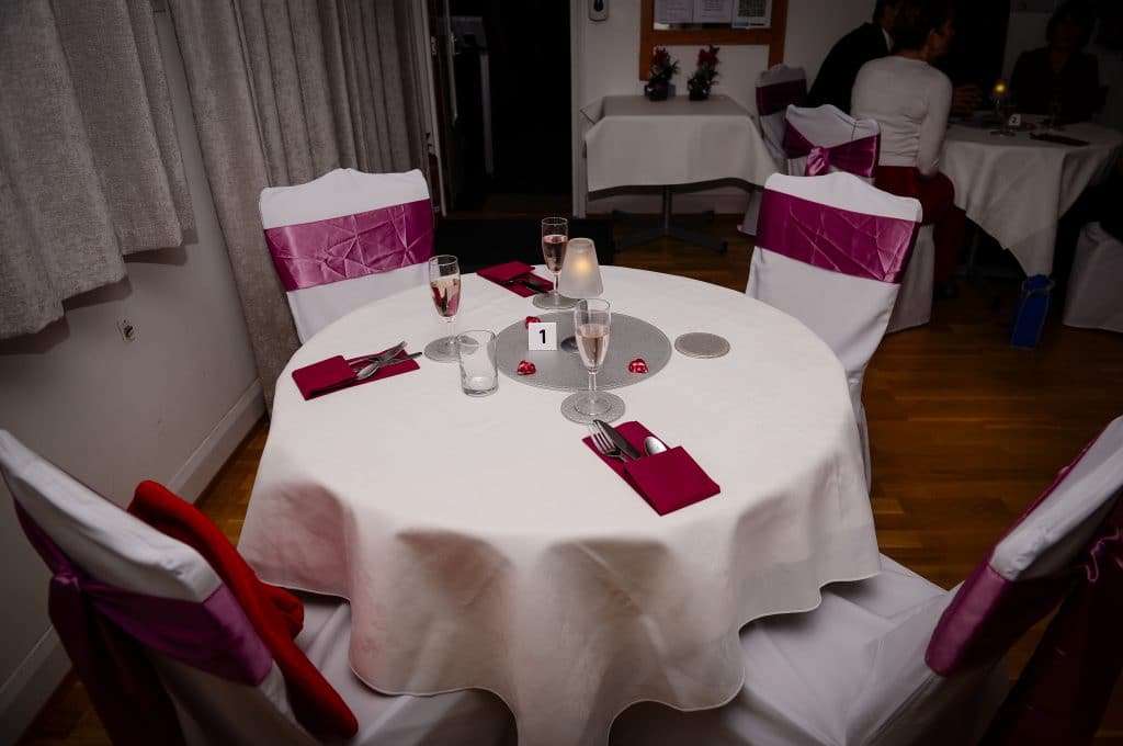 Valentine Dinner & Dance table setup at Learn To Dance in Burnham. A round table with a white tablecloth, burgundy napkin arranged around cutlery with a silver round mat and coasters. Four chairs with white covers and mauve chair sashes.