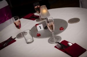 Close up of the table set up at our Valentine Dinner & Dance. White table cloth, silver placemat and coasters. Three champagne flutes filled with rose prosecco. A candle set in the centre