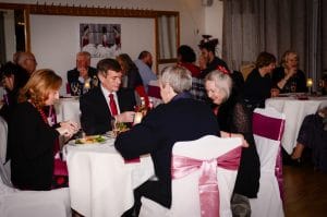 Our Valentine Dinner & Dance in full swing.  Adults sat in tables of four eating dinner.  White covered tables and chairs.  Chairs have mauve satin sashes